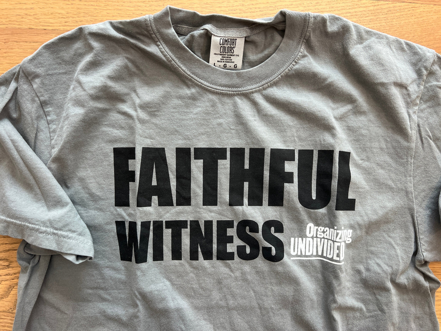 A heather gray short-sleeve t-shirt laid flat on a light wood surface, featuring the words 'FAITHFUL WITNESS' in large black letters and a smaller white graphic reading 'Organizing Undivided' on the chest.
