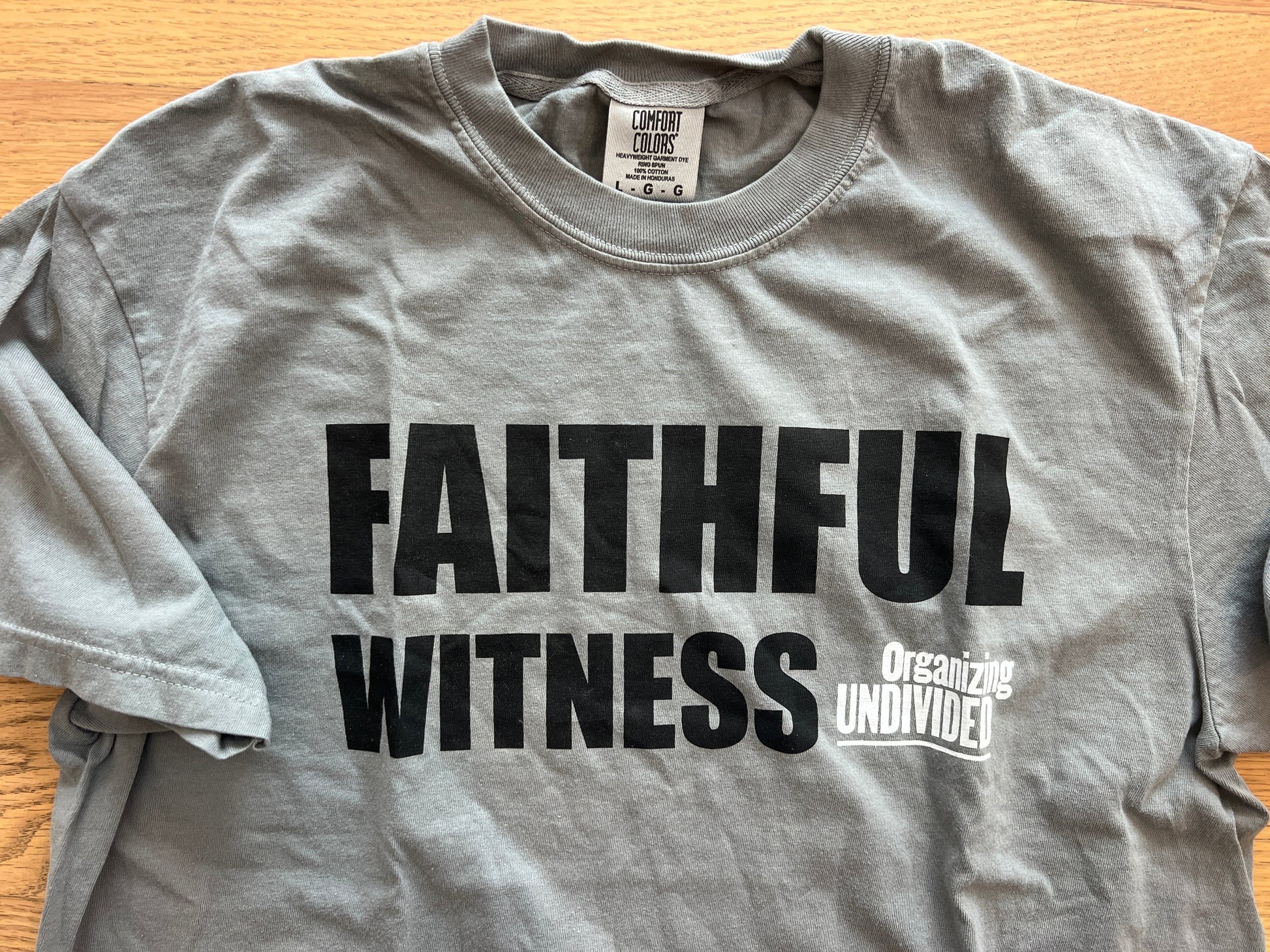A heather gray short-sleeve t-shirt laid flat on a light wood surface, featuring the words 'FAITHFUL WITNESS' in large black letters and a smaller white graphic reading 'Organizing Undivided' on the chest.